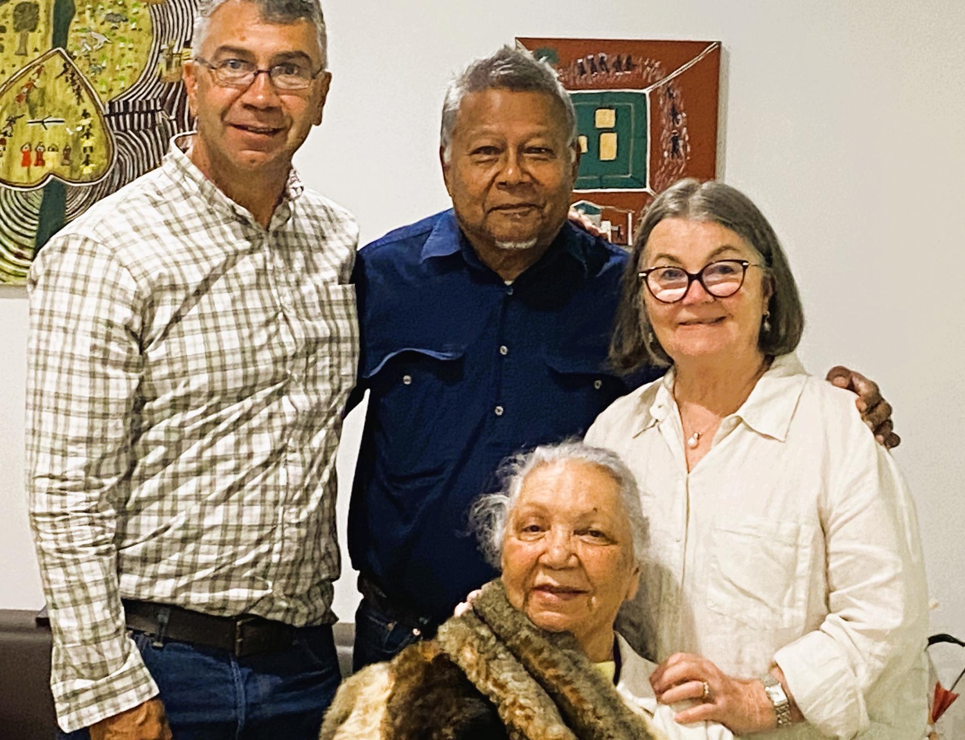 Dr Matilda House-Williams OAM seated with three colleagues at a celebratory gathering, smiling together indoors with artworks displayed on the wall behind them.