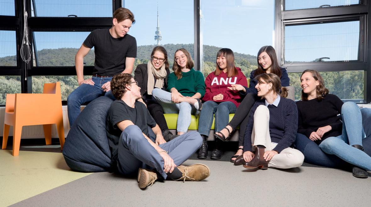 A group of ANU students socialising together in a campus common area, with Black Mountain Tower visible through the windows behind them.