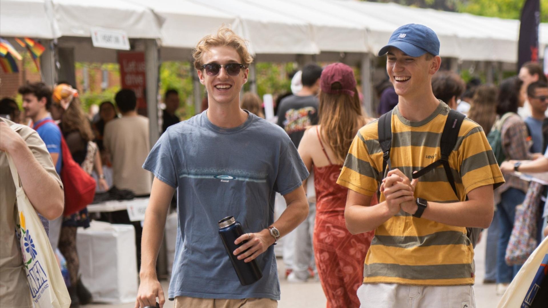 Two students smiling and walking through a busy ANU campus market, with stalls and other students in the background.