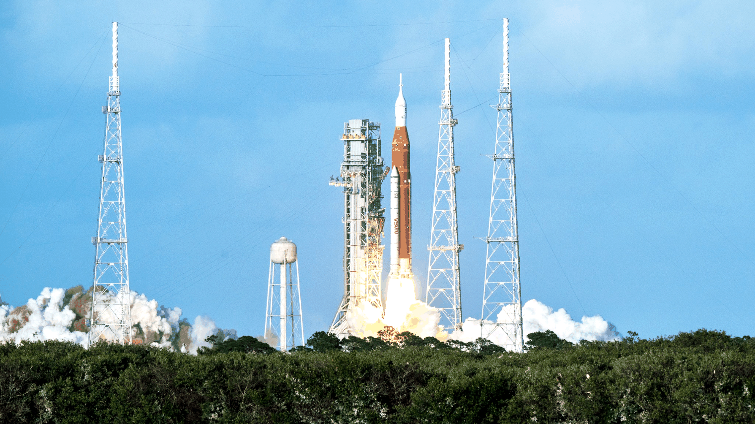 Crowds watch and film the Artemis II rocket launch as it lifts off and arcs through a clear blue sky, leaving a bright smoke trail.