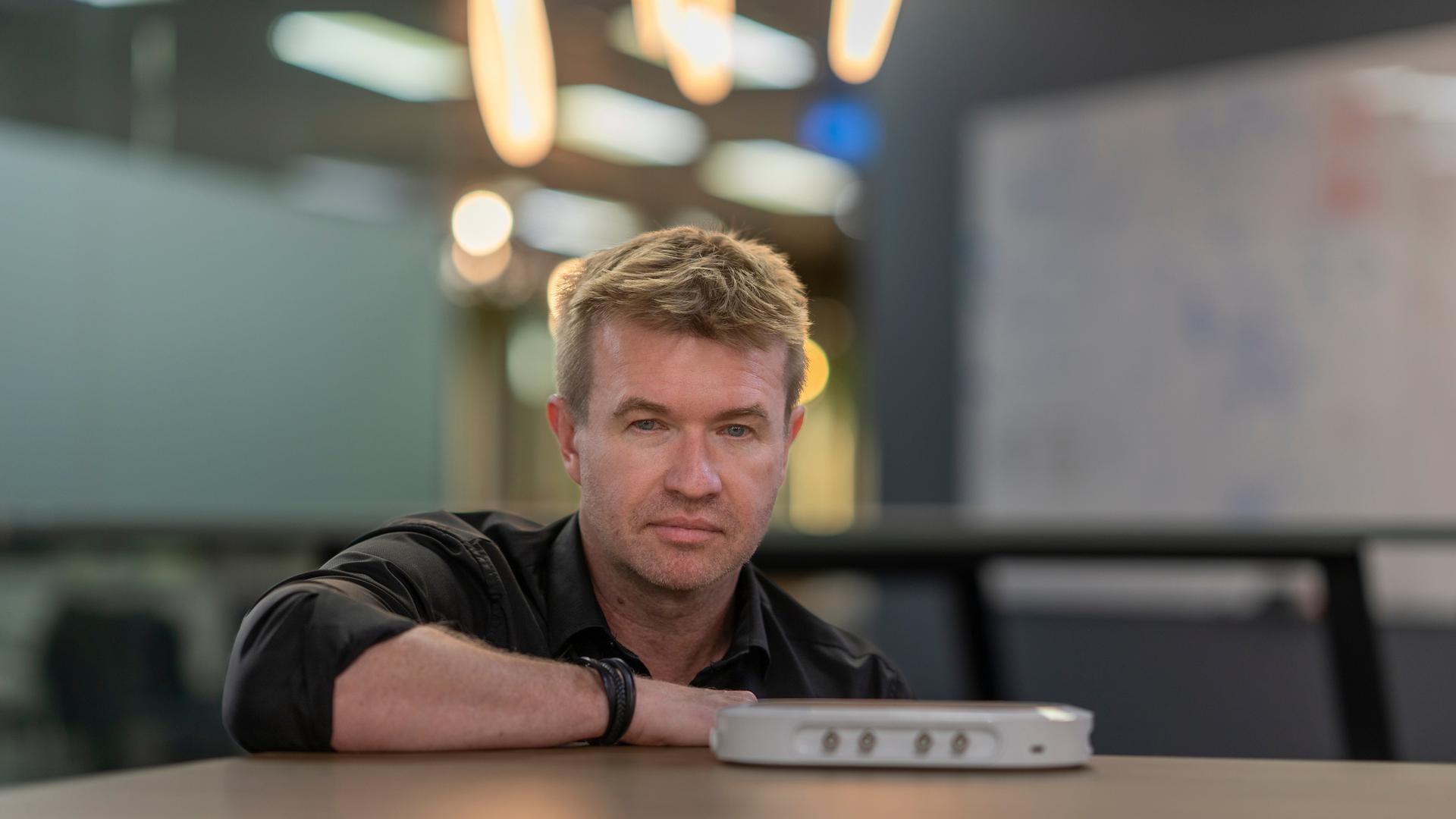 Professor Daniel Shaddock leans on a table, looking directly at the camera, with a small white electronic device placed in front of him. The background is softly blurred with warm overhead lights in a modern indoor setting.