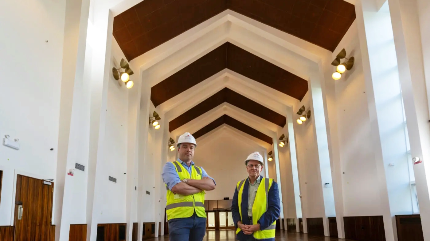 Two construction workers in high-visibility vests and hard hats standing inside a large hall with a vaulted wooden ceiling.