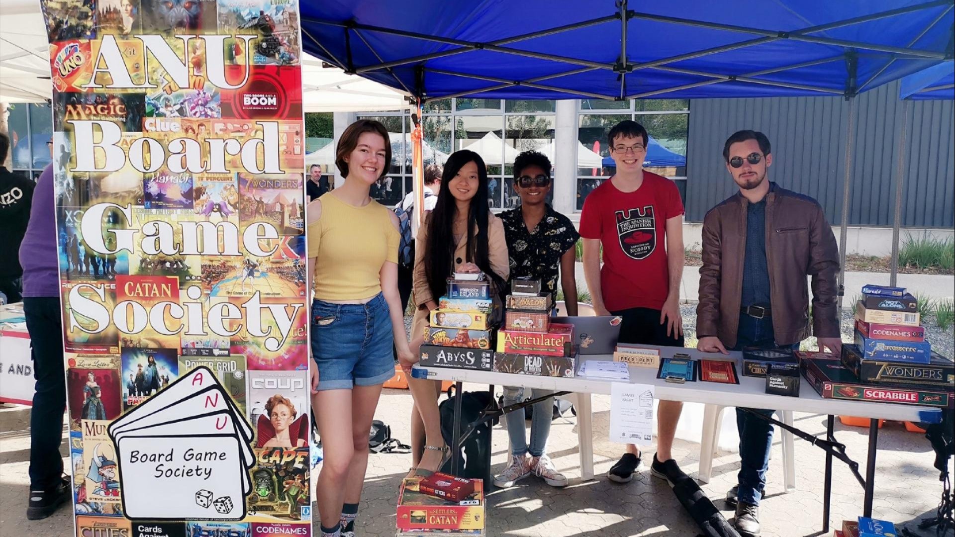 Five people stand behind a table of board games under a blue canopy next to an &ldquo;ANU Board Game Society&rdquo; sign at an outdoor stall.