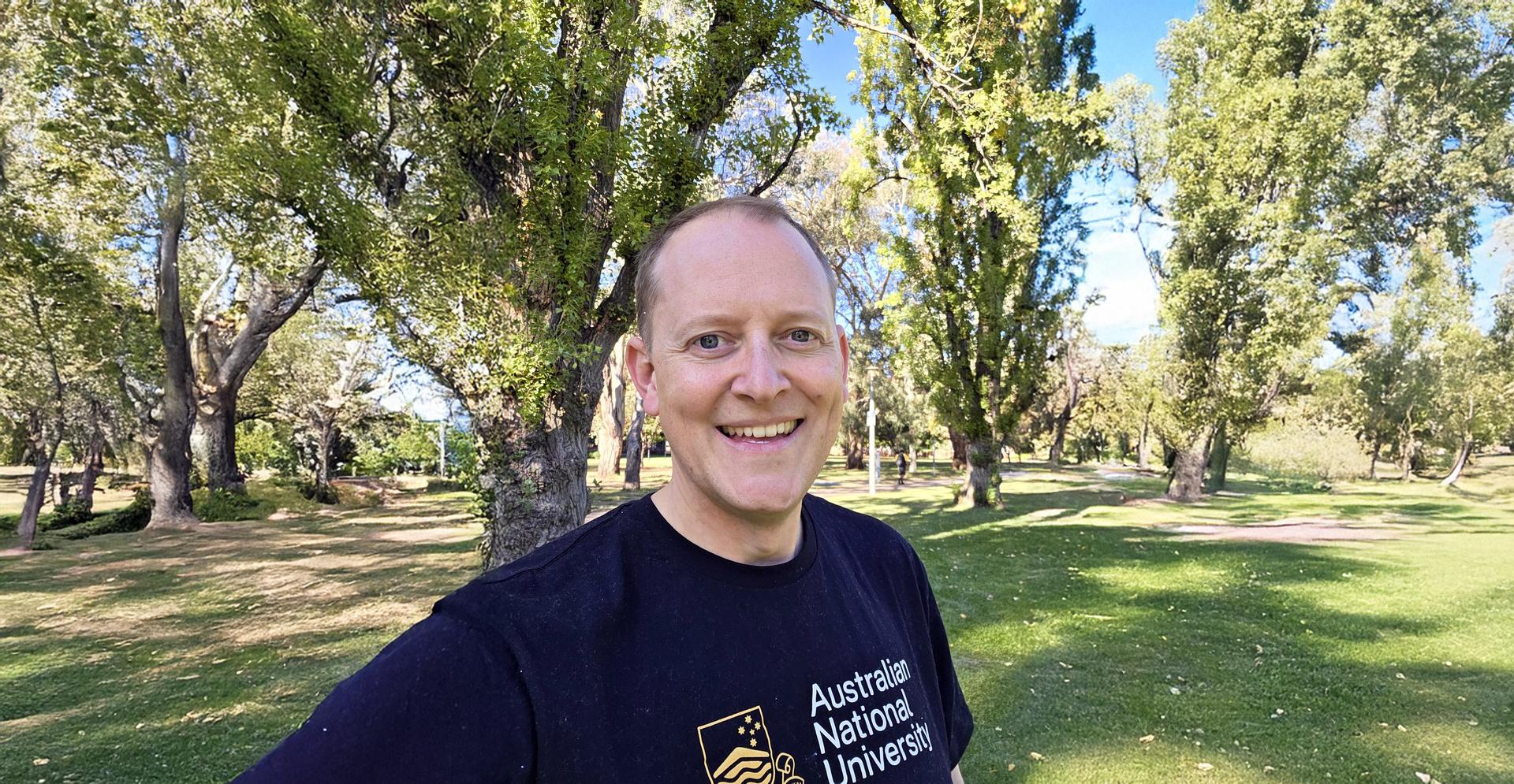 Selfie of Ant Bagshaw smiling in a black tshirt, standing on Campus.