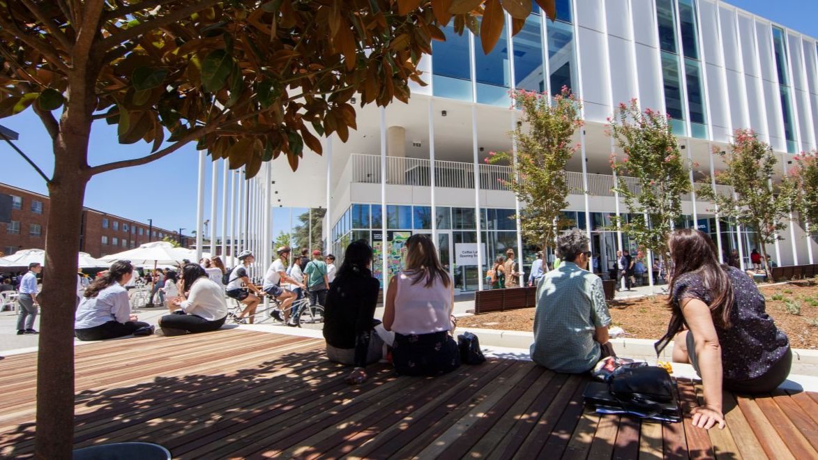 Students relaxing and socialising on a timber deck outside a modern campus building on a sunny day.