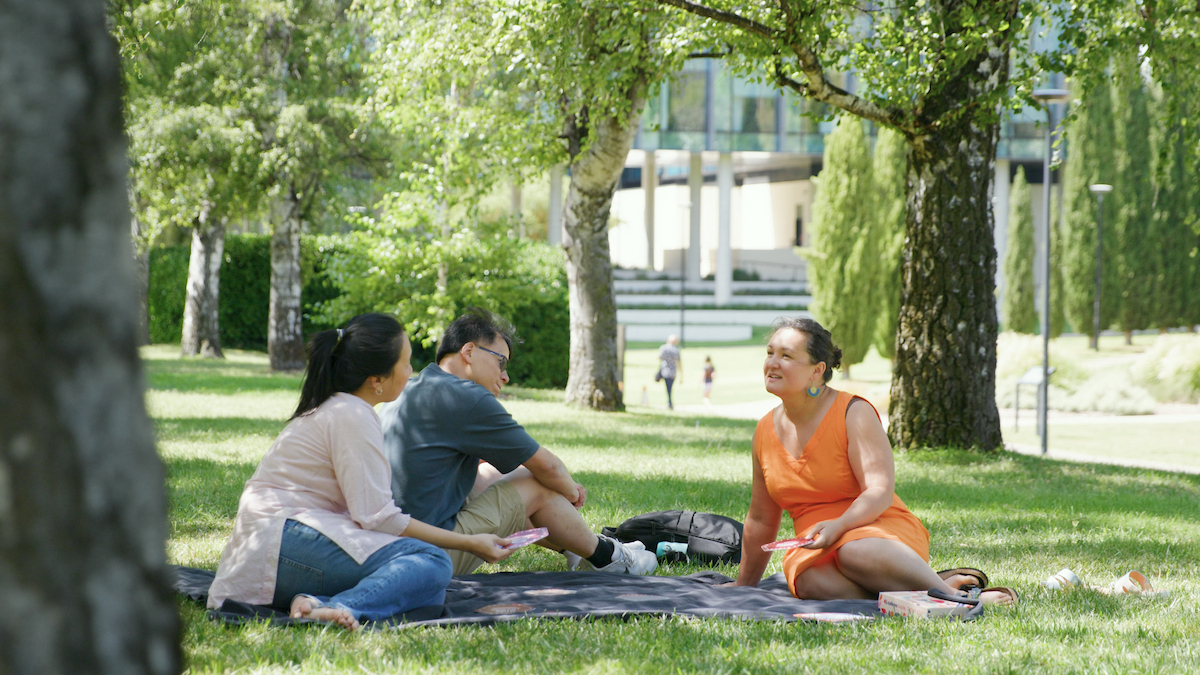 Three people sitting on a picnic blanket under trees on campus, talking and relaxing together outdoors.