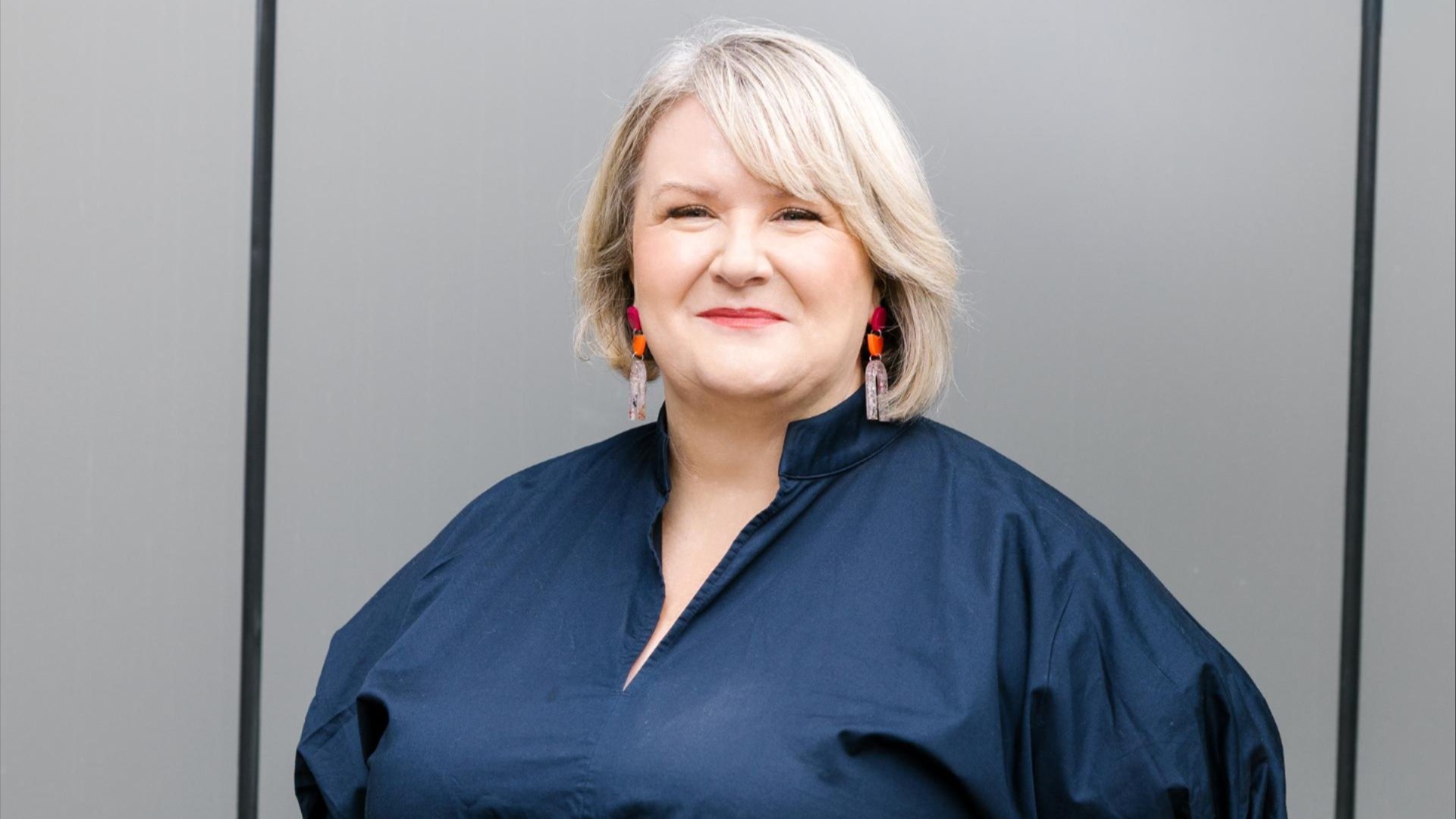 Portrait of Professor Janine O&rsquo;Flynnwith short blonde hair wearing a navy blouse and red earrings against a grey background.