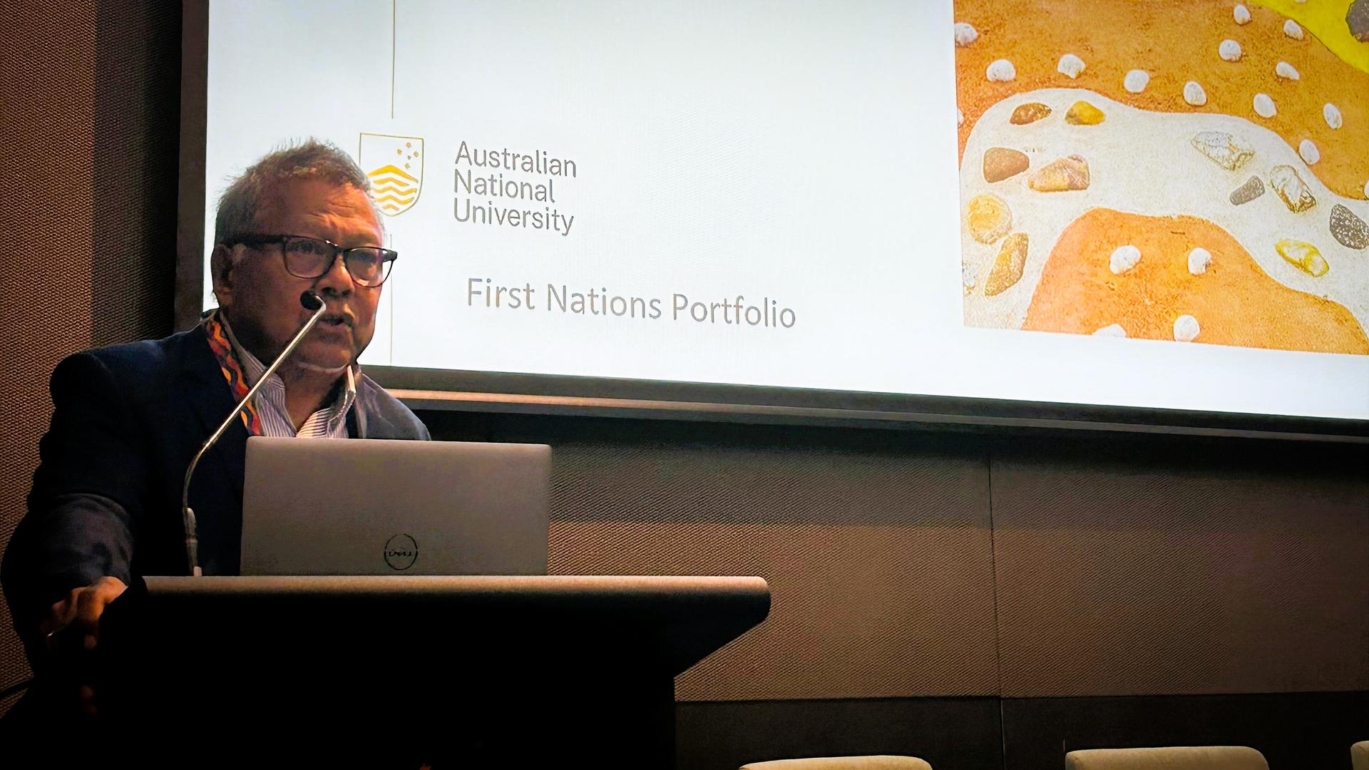 Professor Peter Yu speaks at a podium during the Stronger Smarter Together Summit in Brisbane, with an ��������First Nations Portfolio presentation displayed on screen behind him.