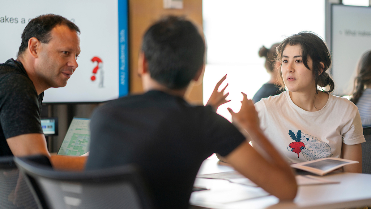 Three people seated at a table in discussion, one man gesturing as she speaks.