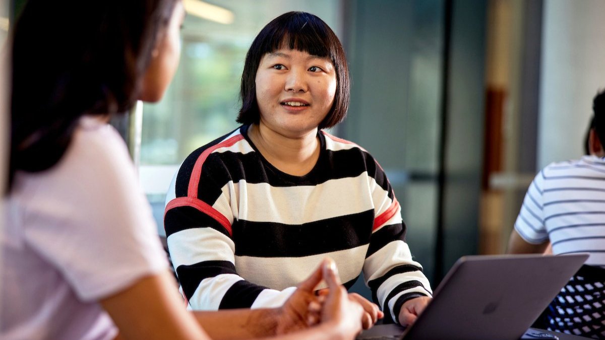 Person in striped sweater speaking with others at a table with a laptop.