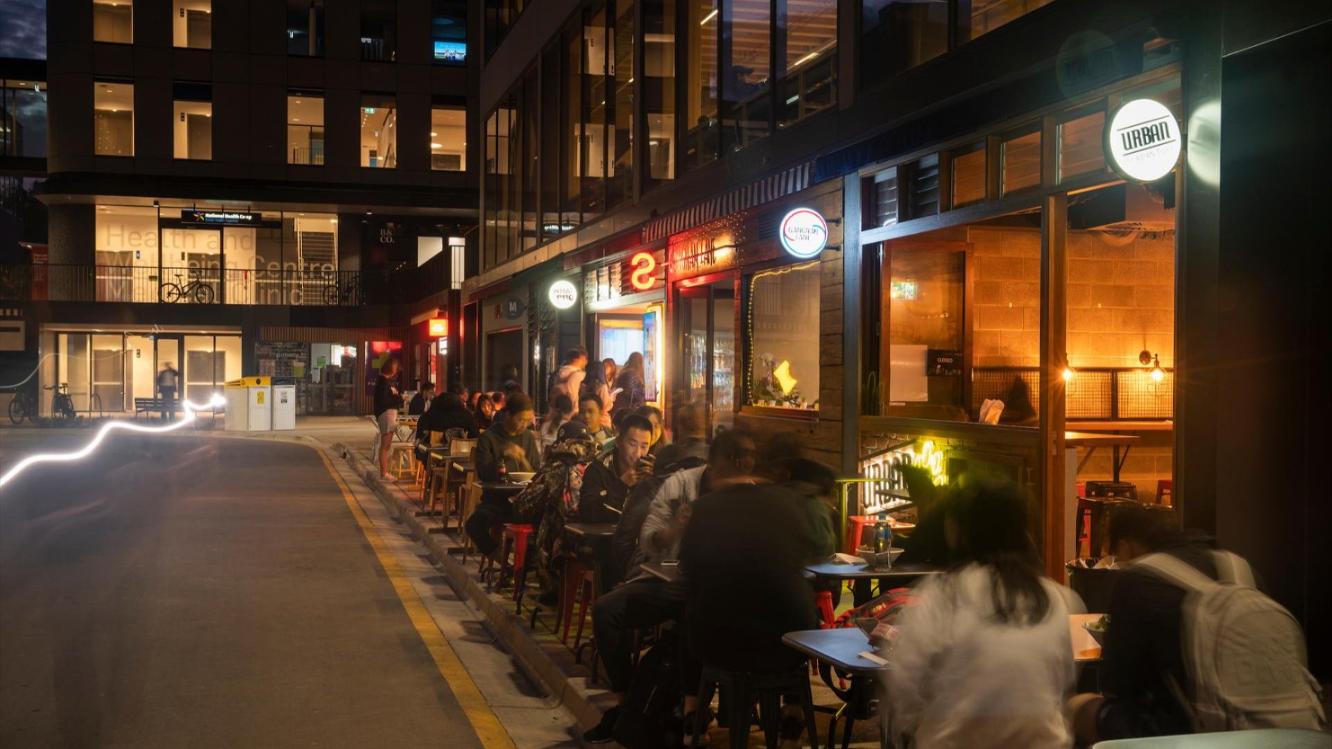 Students and diners at outdoor tables along a busy Joplin Lane at the ��������Acton campus at night, with cafes and restaurants lit up behind them.