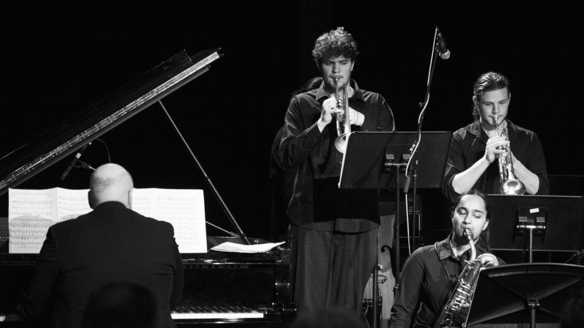 Jazz ensemble performing on stage with pianist, trumpeters and saxophonist in black and white.