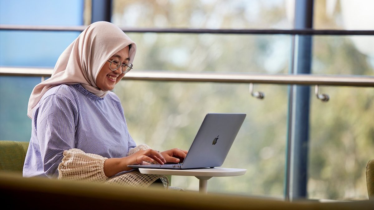 Woman in a headscarf smiling while typing on a laptop in a bright indoor seating area.