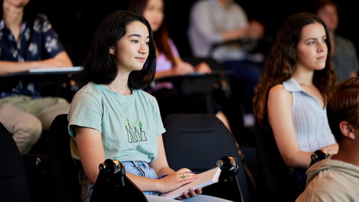 Students seated in a lecture theatre, listening attentively, one taking notes.