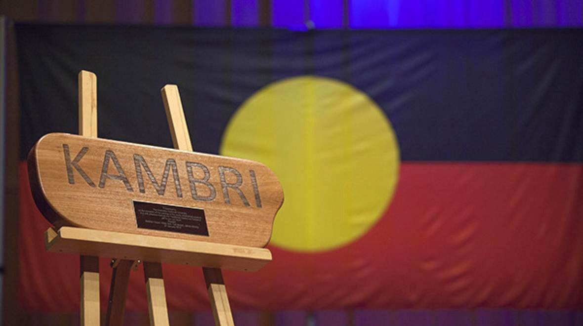 Wooden &ldquo;KAMBRI&rdquo; sign on an easel in front of an Aboriginal flag backdrop.