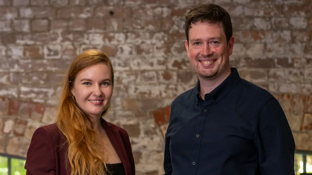 Syenta co-founders Dr Jekaterina Viktorova and Ben Wilkinson standing together in front of a brick wall. 