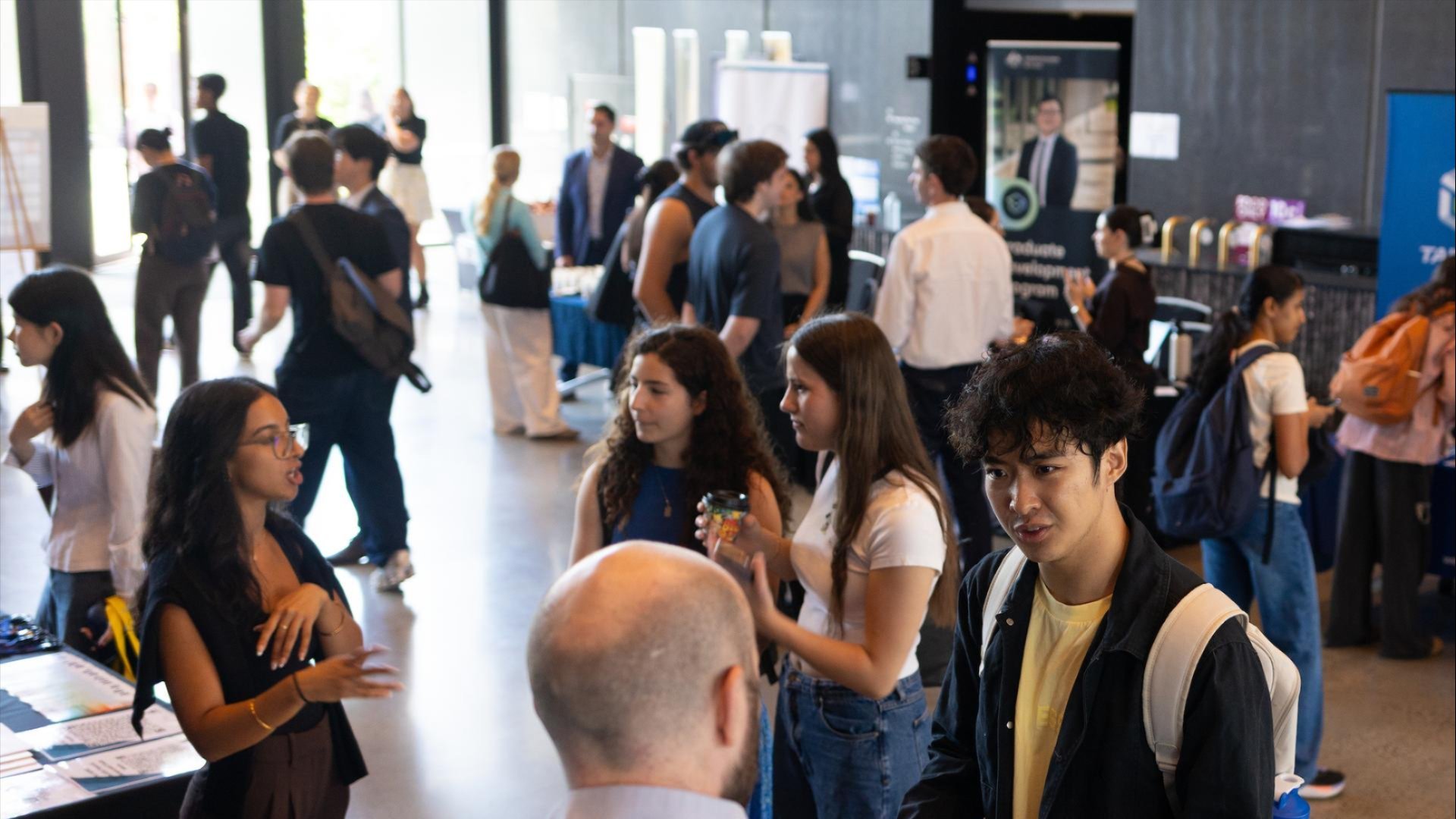 Students chatting with organisation representatives at an ANU fair, with stalls and displays set up inside a busy campus venue.