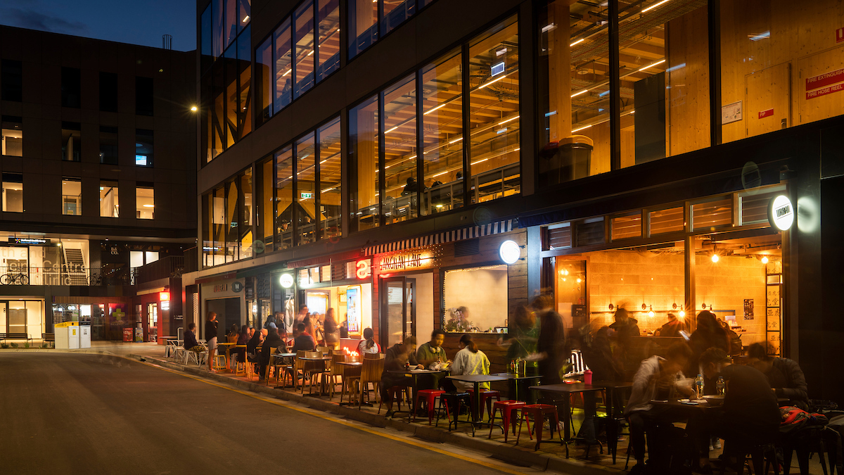 Outdoor dining along a lit street at night beside a modern glass-fronted building, with people seated at tables.