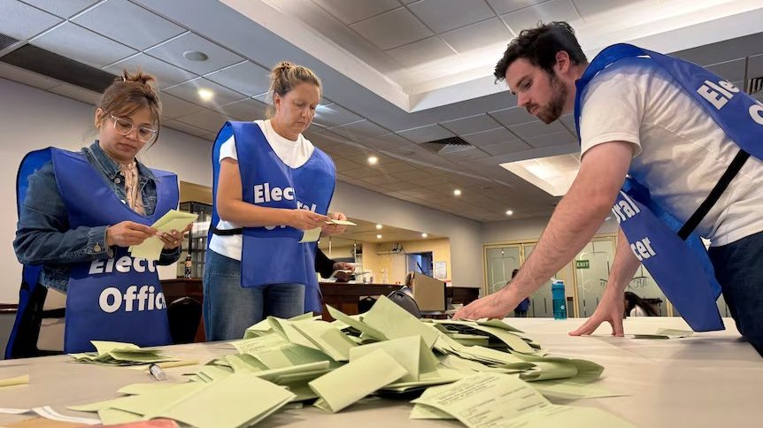 Election workers sorting ballot papers on a table in an indoor counting room.
