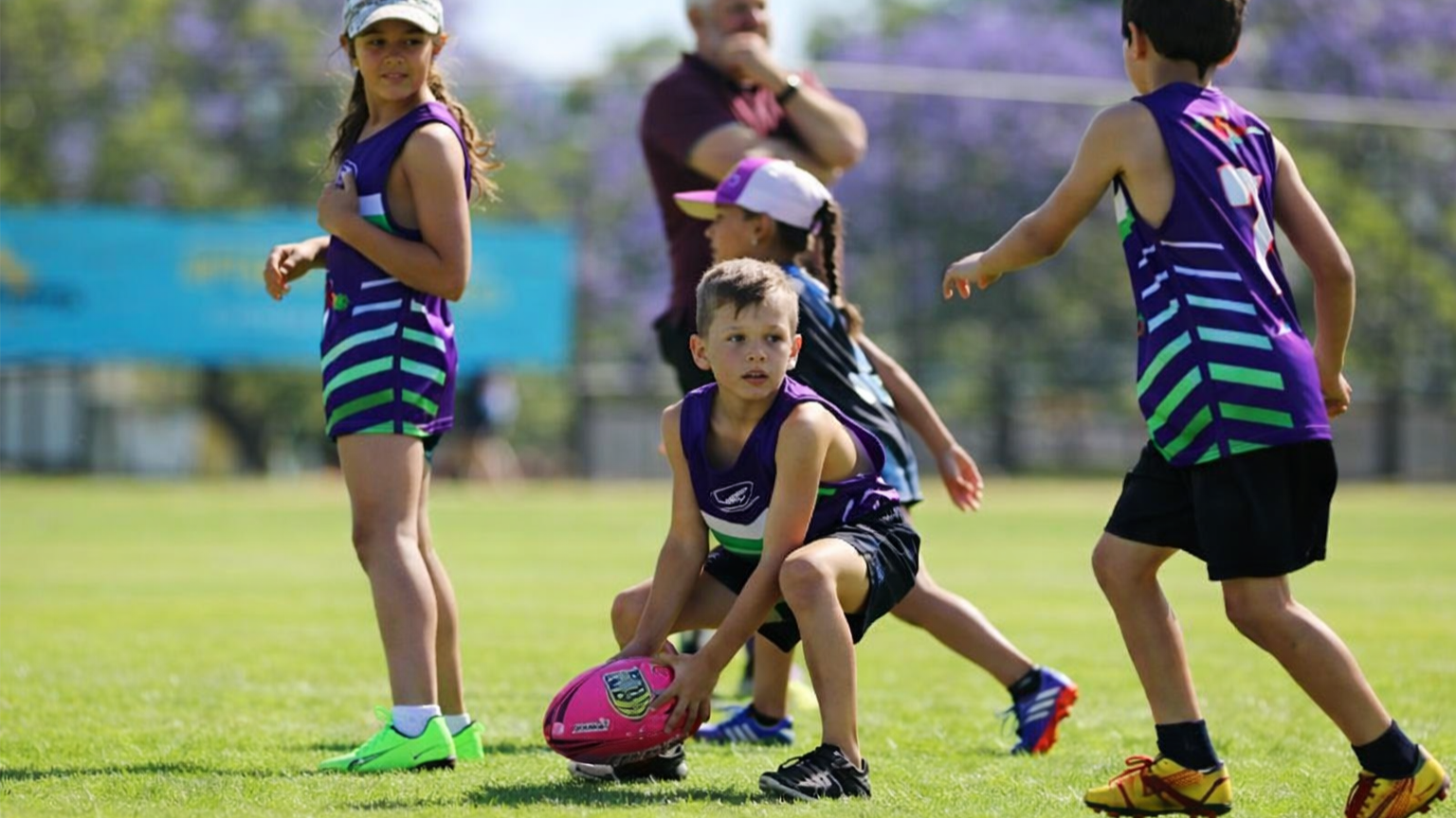Children in purple sports uniforms play with a football on a grassy field while a coach watches in the background.