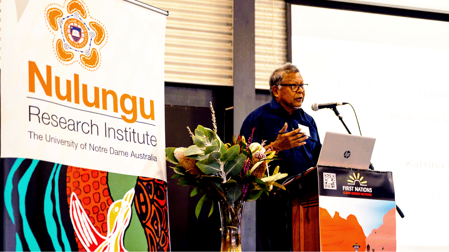 Speaker at podium with microphone beside &ldquo;Nulungu ���˸�����ҹӰԺ Institute&rdquo; banner, delivering a talk.