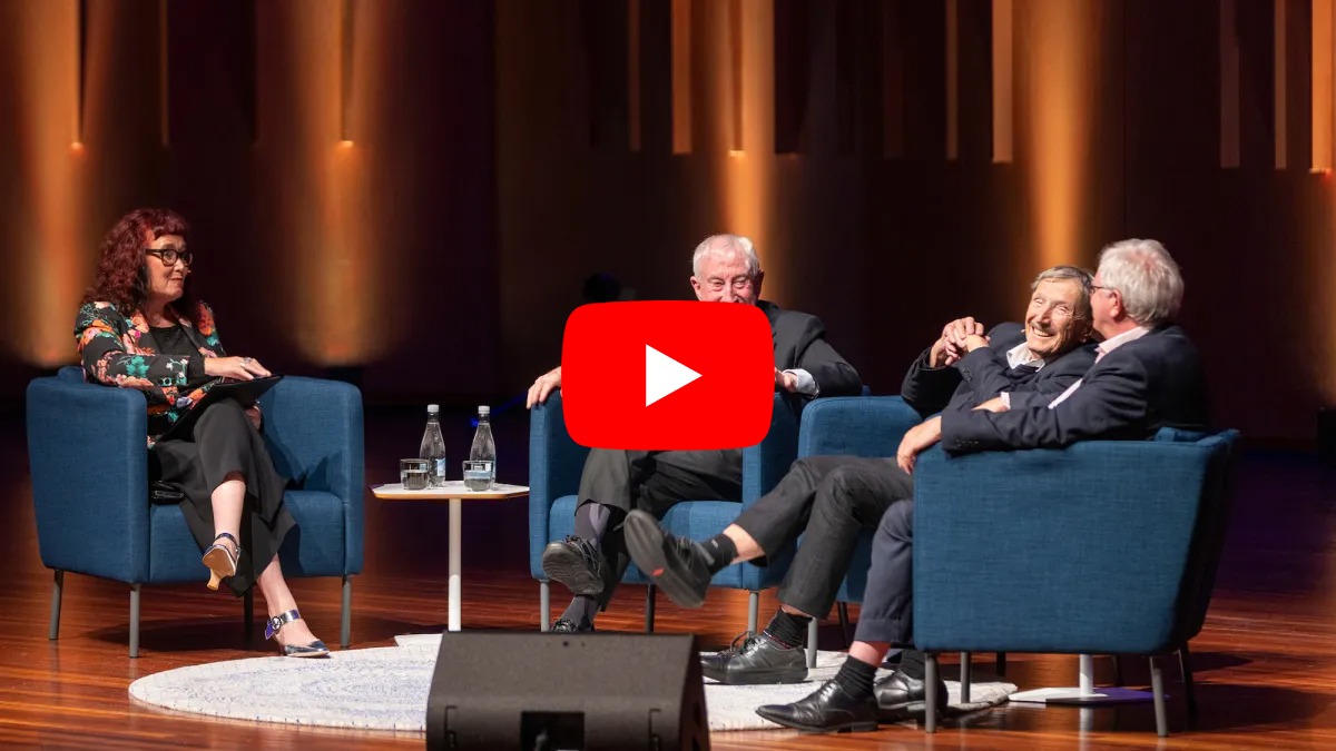 Panel discussion with four speakers seated on stage in armchairs, with a video play icon overlay.