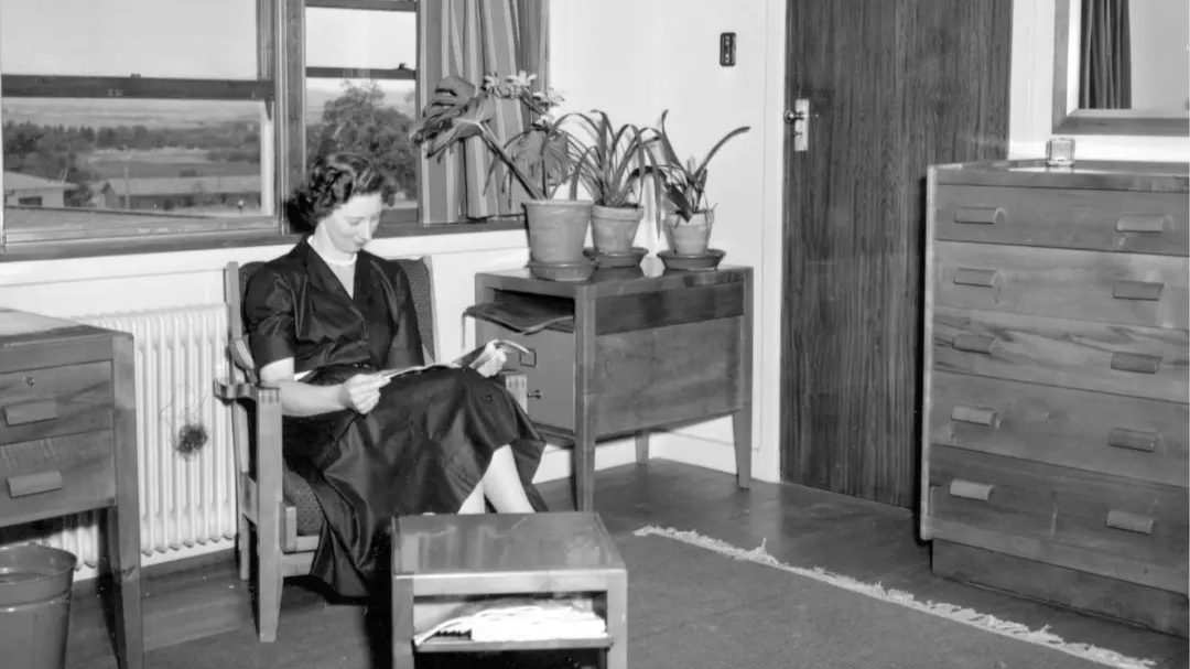 A woman reads in a Fred Ward-designed armchair inside a University House room, circa 1954, surrounded by original Ward furniture including a writing desk and chest of drawers.