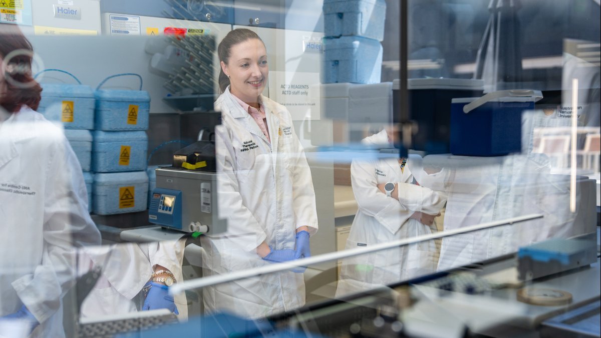 Scientists in lab coats and gloves working in a laboratory, viewed through glass with equipment in the foreground.