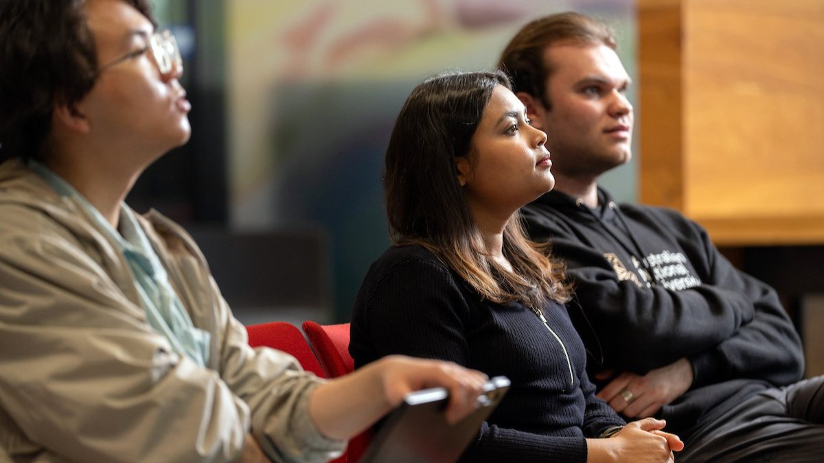 Three people seated and listening attentively, one holding a pen and notebook.