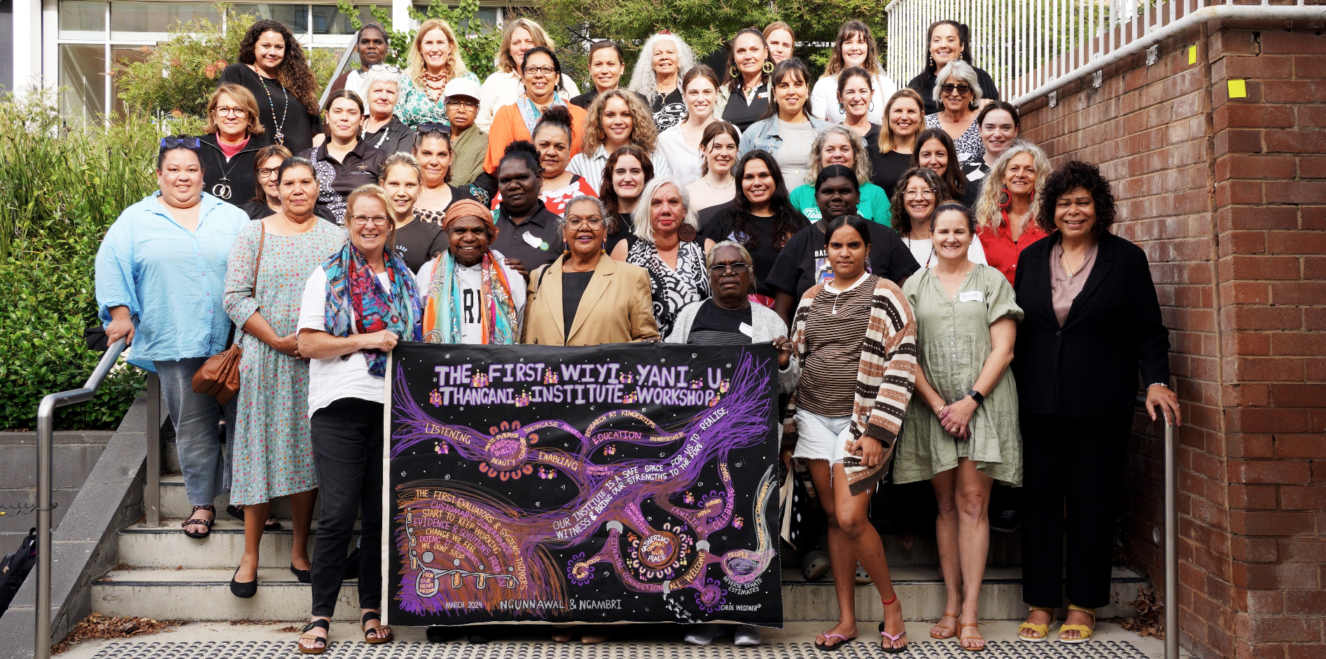Large group of people gathered on outdoor steps holding a colourful workshop banner.
