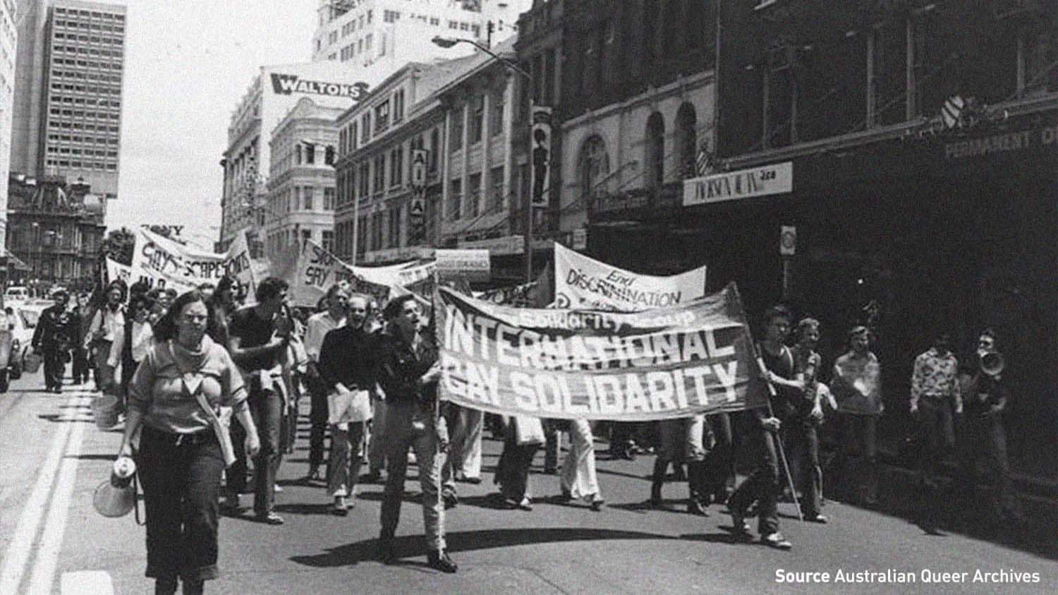 Black-and-white photo of a street march for LGBTQIA+ rights, with people carrying banners through a city street.