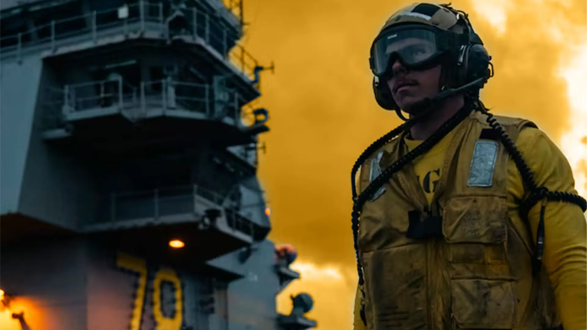U.S. Navy flight deck crew member in a yellow vest and helmet holding illuminated wands, standing on an aircraft carrier deck at dusk with other crew and the ship&rsquo;s island structure in the background.