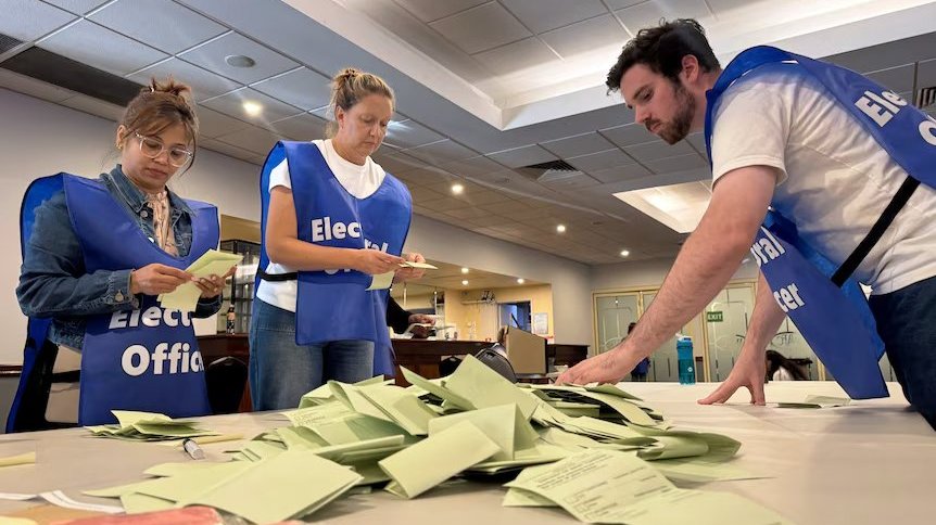 Election workers sorting ballot papers on a table in an indoor counting room.