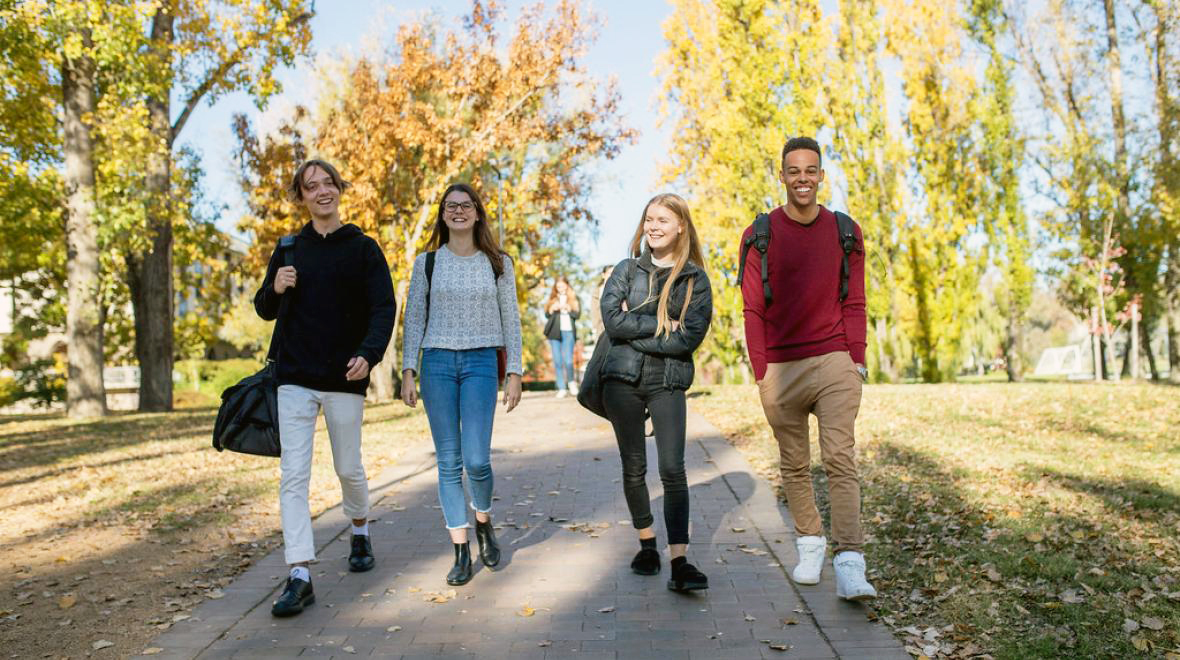 Four students with backpacks walking and smiling along a tree-lined path with autumn foliage.
