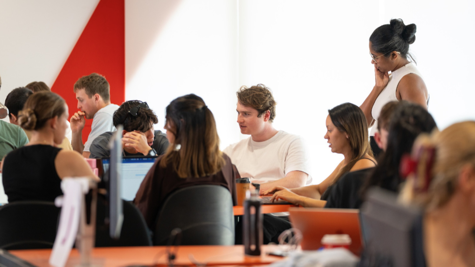 Students working together in a collaborative classroom space, with a staff member standing nearby and others seated at desks using laptops.