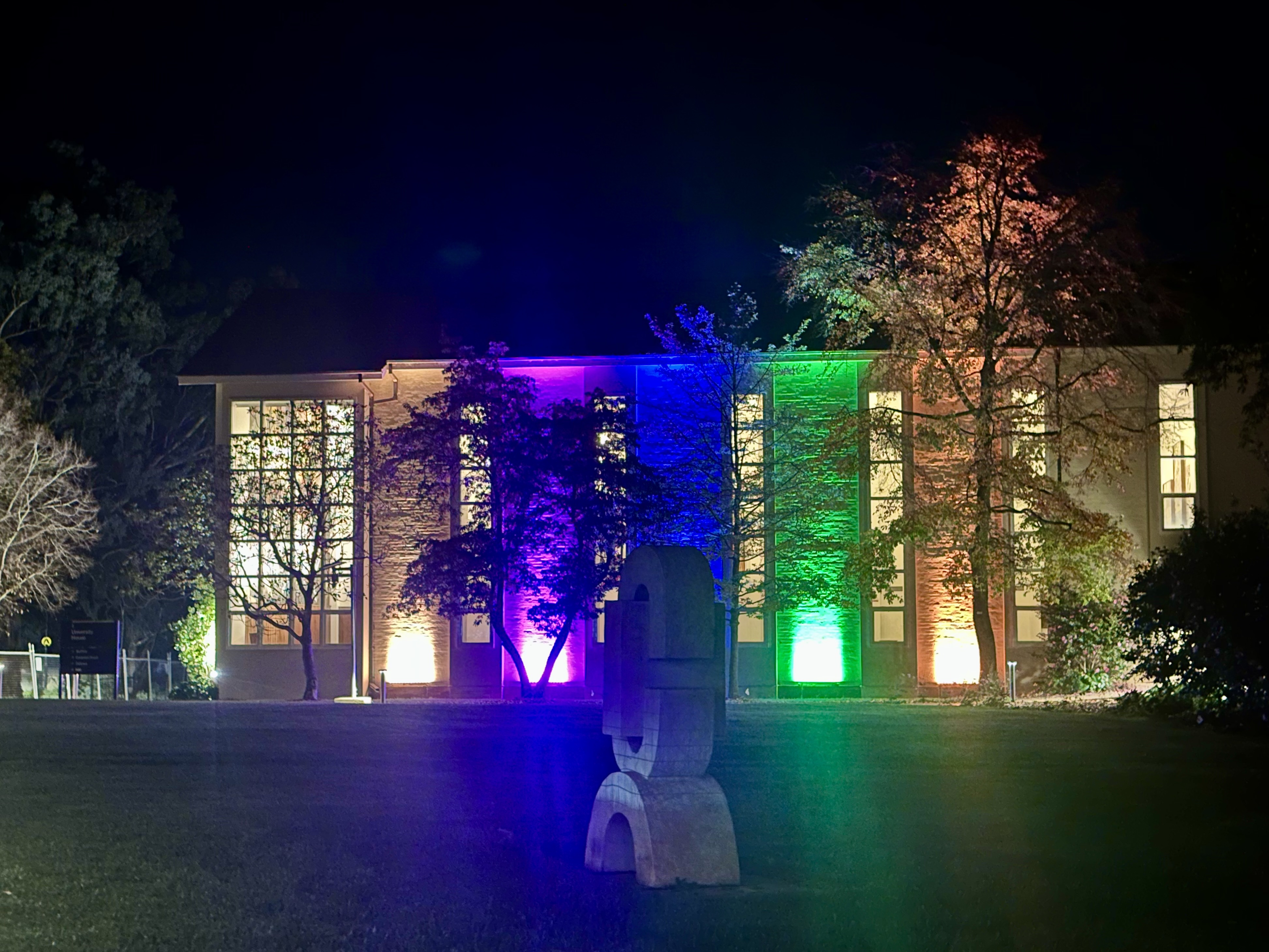 University House at ������Ƶlogo lit in rainbow colours at night, with illuminated trees and a sculpture in the foreground.