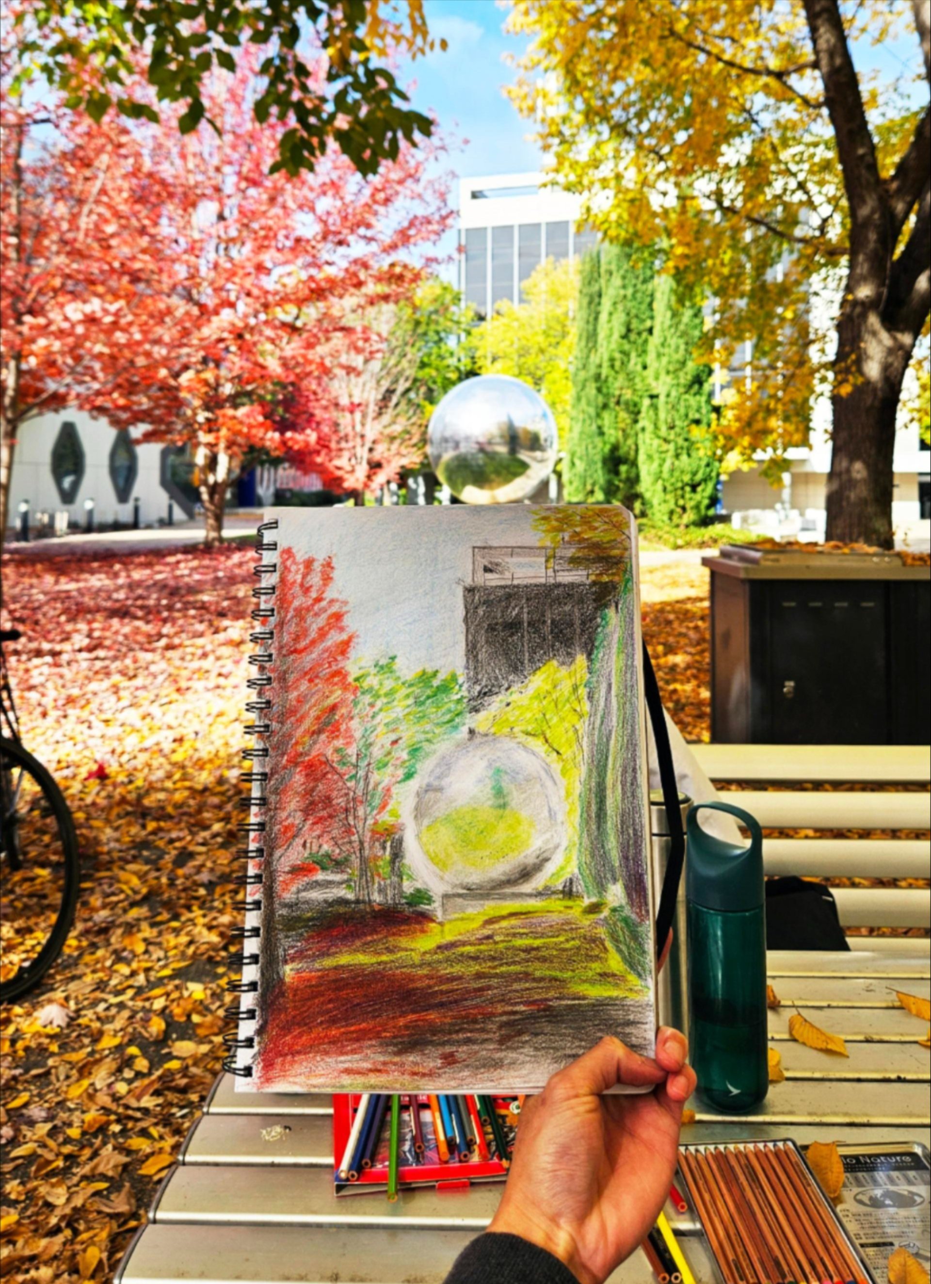 Hand holding a sketchbook drawing of the mirrored sphere amid colourful autumn trees.