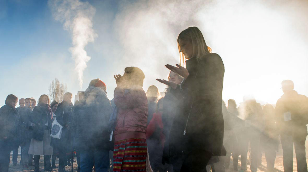 People gathered outdoors during a smoking ceremony, with smoke drifting through the sunlight.