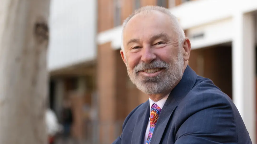 Dr Gordon de Brouwer smiles at the camera outdoors on the ANU campus, wearing a dark suit and colourful patterned tie.