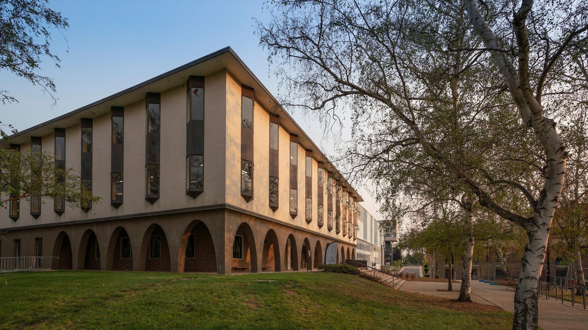 Exterior of Chifley Library at dusk, showing its arched ground-floor colonnade and tall vertical windows, with trees in the foreground.