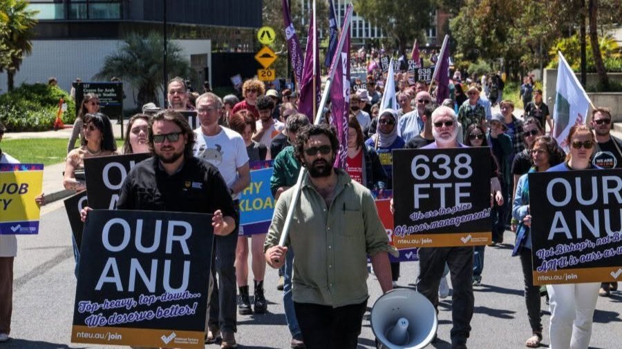 NTEU demonstrators march through the ANU campus carrying union flags and placards, with a person holding a megaphone at the front.