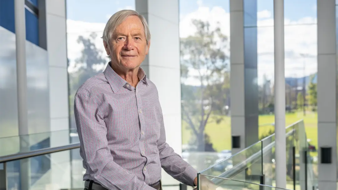 Emeritus Professor Trevor Lamb stands beside a glass balustrade in a bright ��������building.