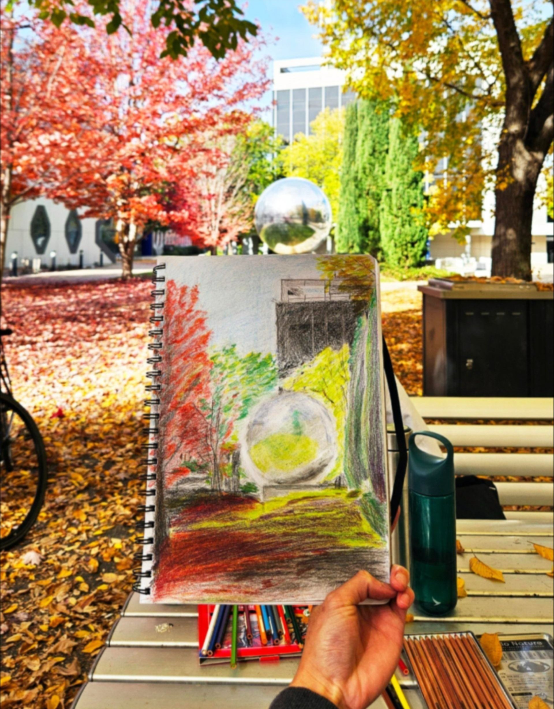 Hand holding a sketchbook drawing of the mirrored sphere amid colourful autumn trees.