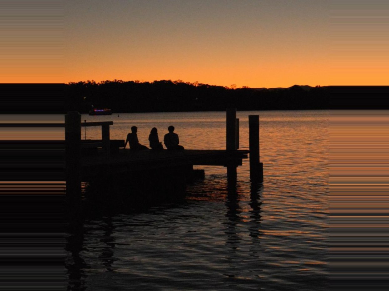 Silhouette of three people sitting on a jetty on Lake Burley Griffin at sunset, with hills in the background.