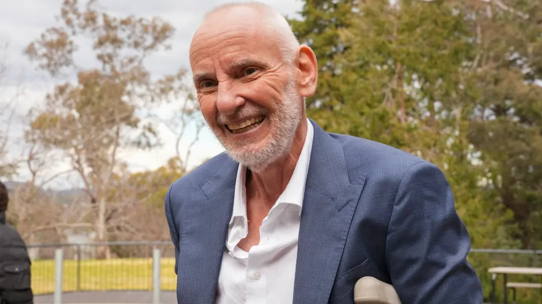 Dr Chris Taylor smiles outdoors, wearing a blue blazer and white shirt, with trees in the background.
