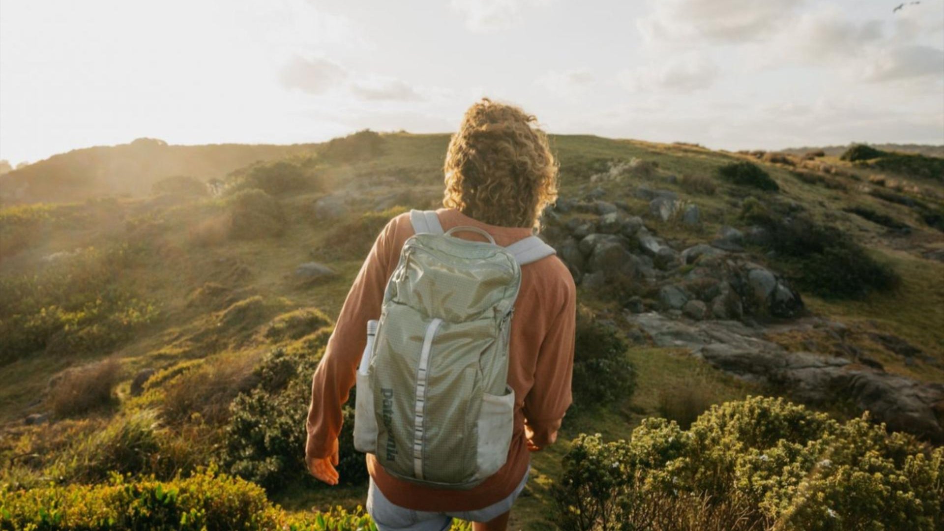 A person wearing a backpack walks along a grassy coastal headland at sunset.
