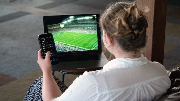 A woman watches a football match on a laptop while holding a smartphone.