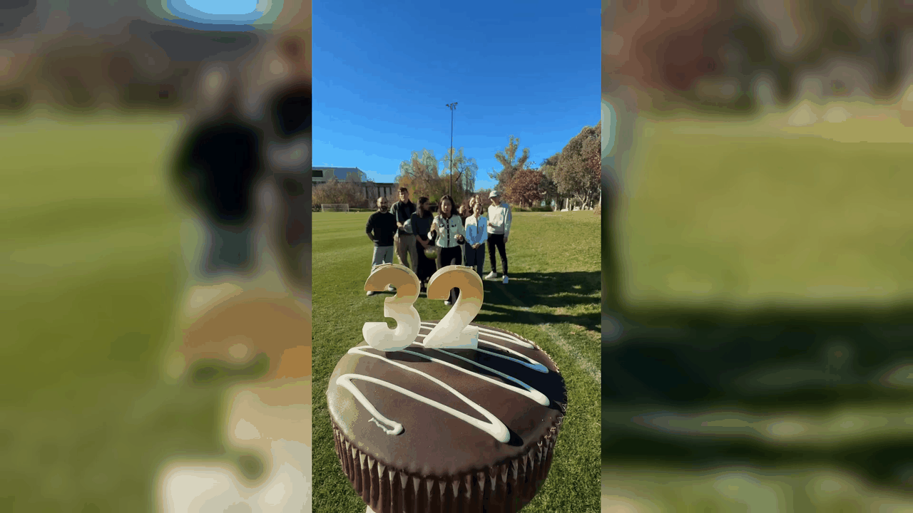 Animated video showing ANU staff and students standing on a campus lawn behind a chocolate cake topped with number candles. The video is presented in a landscape format with the original portrait footage centred against a blurred background.