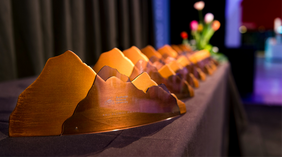 ANU Alumni Award trophies displayed in a row on a table, with the nearest award in focus and the others fading into the background.