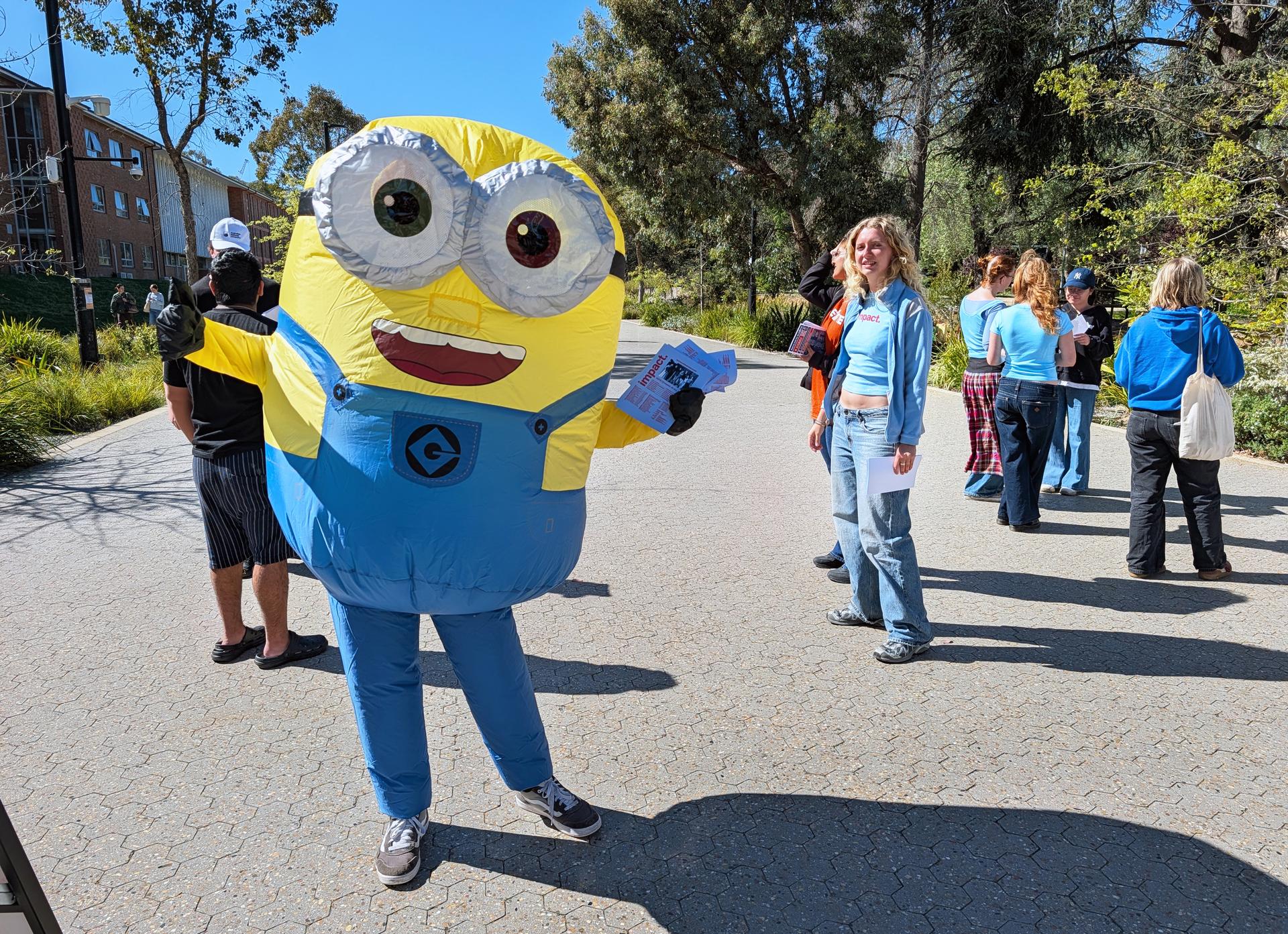Person dressed as a Minion at an outdoor ��������event, with students in the background.